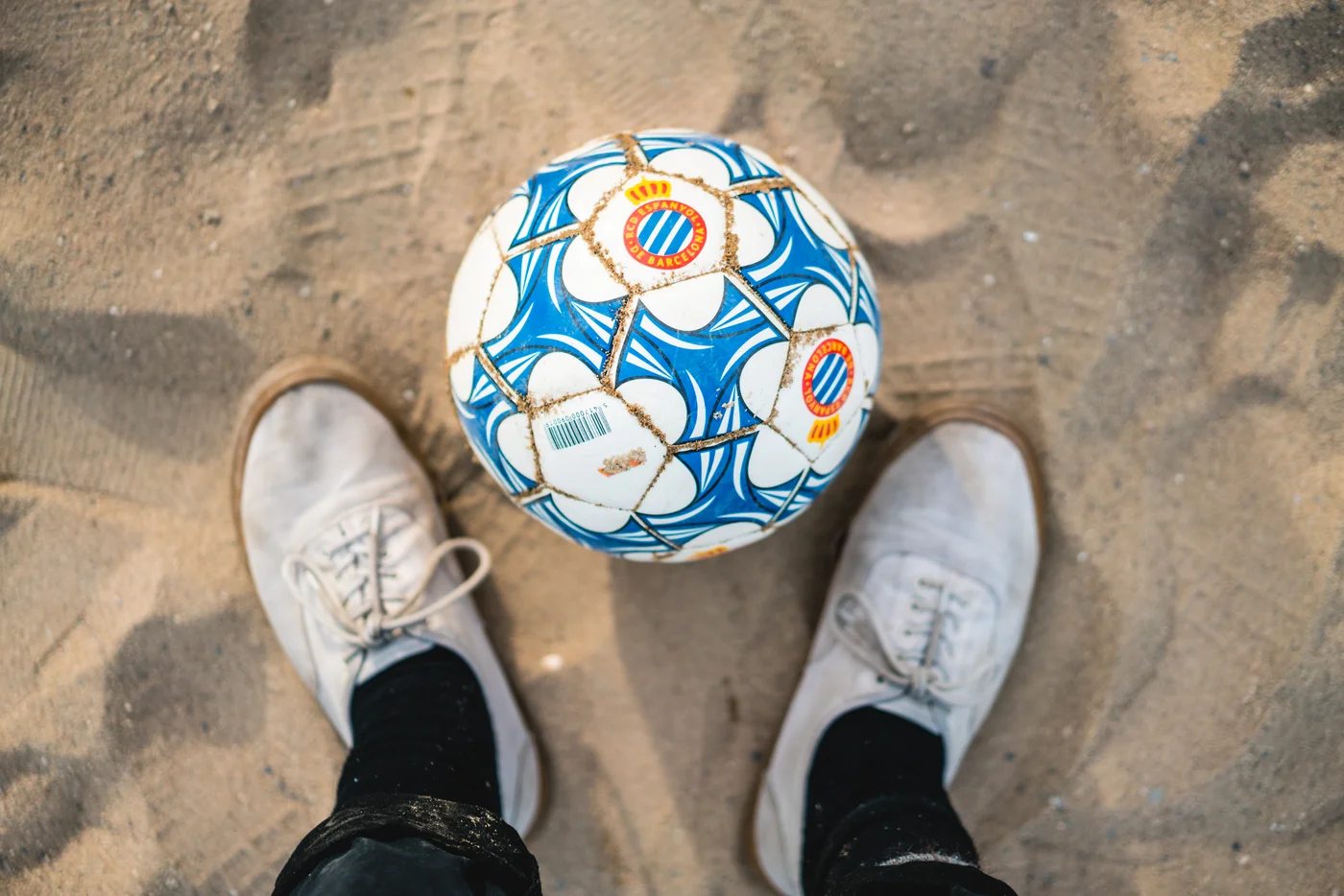blue and white soccer ball on sand with persons white casual sneakers with black socks and black jeans rolled up
