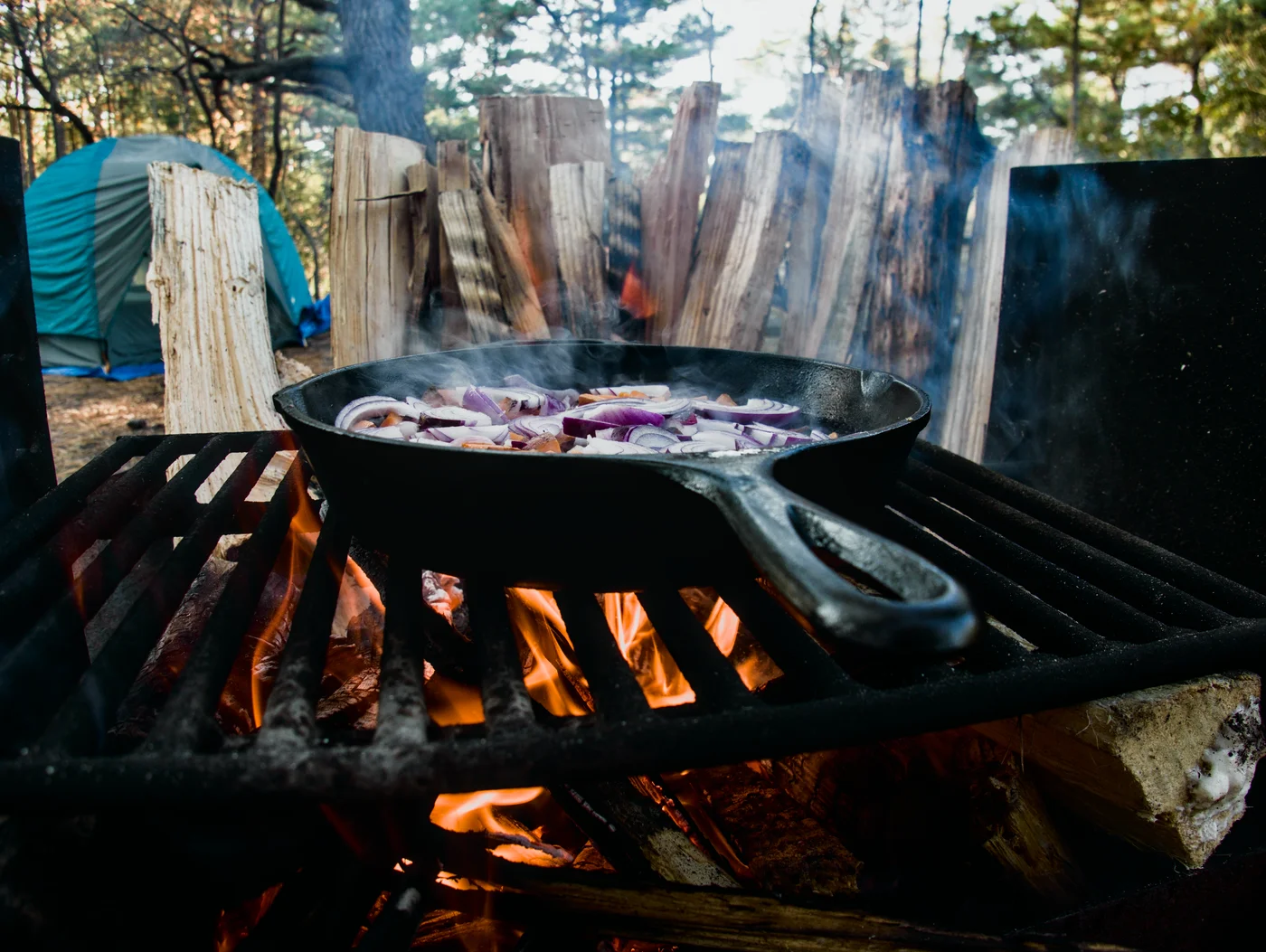Sweet potatoes and red onion cooking in a cast iron pad over a firepit with a grill. Wet firewood is stacked behind it to dry out over the fire
